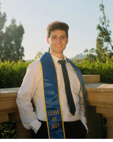 Bassel Lawand smiling on a stone balcony in late-afternoon sun, in a white shirt, a dark tie and a blue UCLA stole that reads Class of 2025.