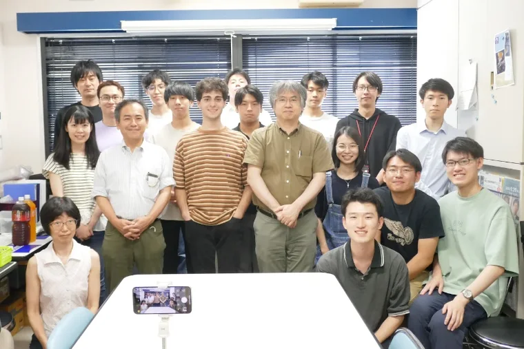 The Ohashi Lab group in their lab room at Tohoku University, about twenty people. Bassel stands centre-left in a striped shirt.