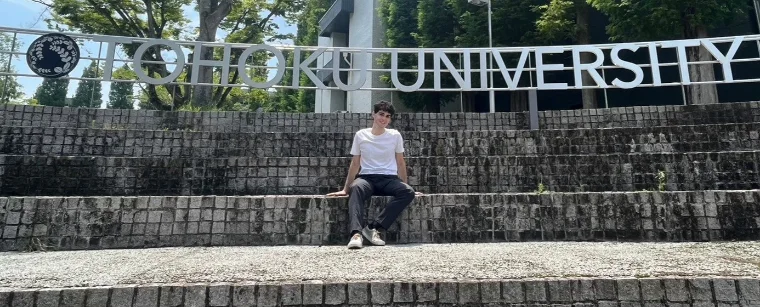 Bassel sitting on wide stone steps below the TOHOKU UNIVERSITY sign, tall trees behind him on a bright summer day.