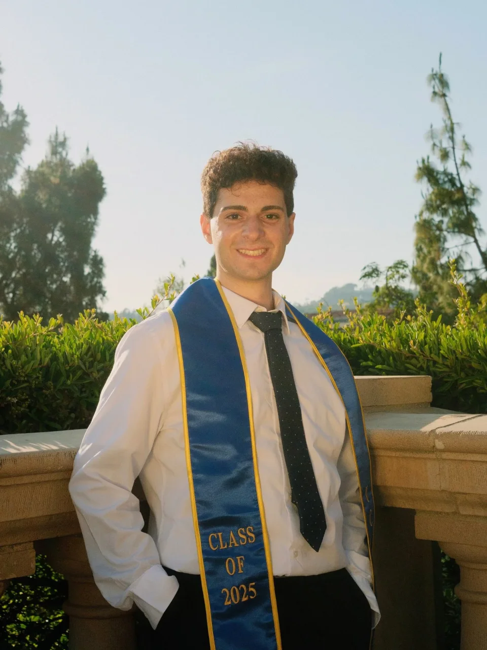 Bassel Lawand smiling outdoors in a white shirt and dark tie, wearing his UCLA Class of 2025 graduation stole.